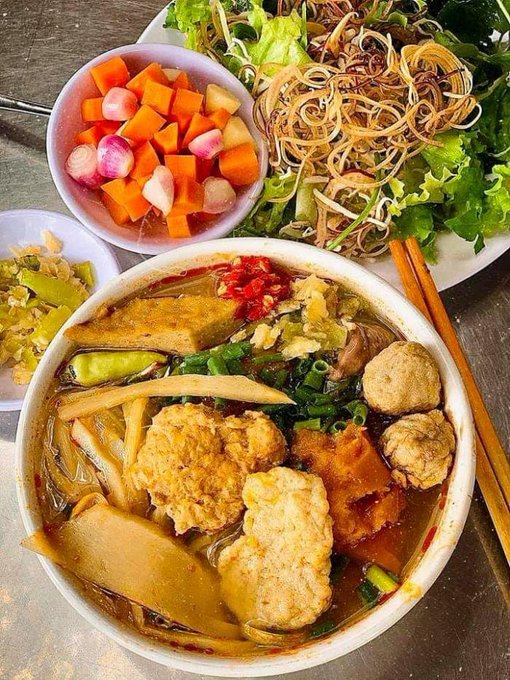 An overhead shot of a Vietnamese meal featuring a bowl of bún chả or similar soup with fish cakes, fried items, and vegetables, accompanied by a plate of pickled vegetables, fresh herbs, and rice noodles, with chopsticks on the side.