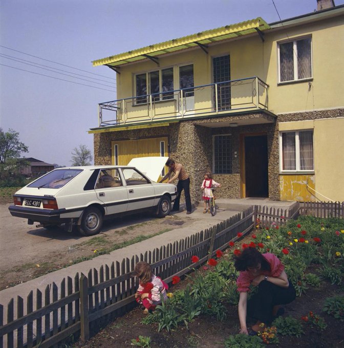 A family tends to their garden and a white hatchback car (license plate '34 C 437') in front of a yellow two-story house with a balcony, as a man works under the car's hood and children play nearby.