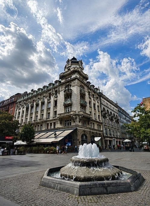 A grand Beaux-Arts style corner building with ornate architecture, situated behind a tiered water fountain in a cobblestone plaza under a dramatic blue sky filled with white clouds.