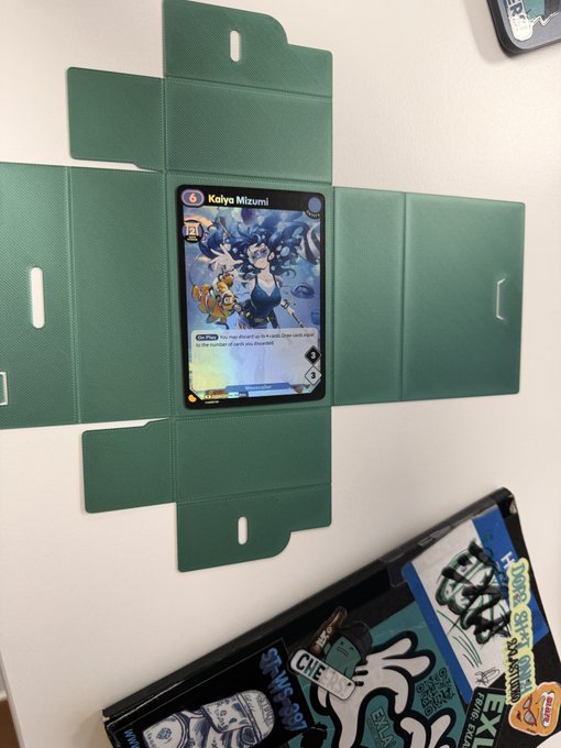 A green Azuki TCG deck box prototype is shown open, displaying a Kaiya Mizumi card in the center, with additional sticker-covered storage items visible to the side. The image showcases the creator's handmade deck box design, relevant to their announcement about producing BEANZ-themed Azuki TCG deck box prototypes.