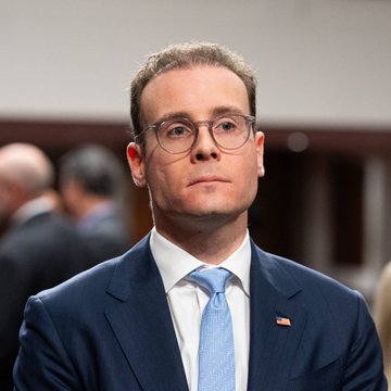 A young man with brown hair and glasses, wearing a dark navy suit, white shirt, and light blue tie with an American flag pin on his lapel, appears to be at a formal governmental proceeding. The image relates to the post as it likely depicts the CFTC Chair Selig being referenced in the headline about the agency's readiness to proceed with regulations regarding digital assets.