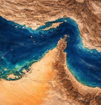 A satellite or aerial view showing a narrow body of turquoise and deep blue water separating arid, brown desert landscapes, likely depicting the Strait of Hormuz.