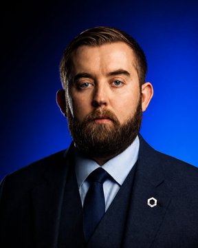 A portrait of a bearded man wearing a dark suit and tie against a blue background, featuring a small hexagonal pin on his lapel.