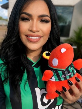 A woman wearing a green and black BC.GAME jersey smiles while holding a red octopus plush toy wearing a matching BC.GAME shirt, directly promoting the brand's rewards program and soccer partnership mentioned in the post.