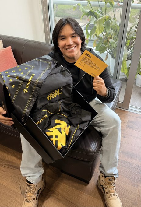 A smiling young man with dark hair sits on a couch holding a yellow card and a black box containing Binance-branded merchandise including a hoodie and other items.