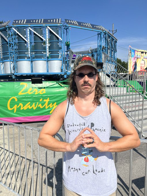 A man with long hair, mustache, and sunglasses stands in front of a 'Zero Gravity' carnival ride, wearing a handwritten tank top reading 'Graduated University of Phoenix Magna Cum Laude'.