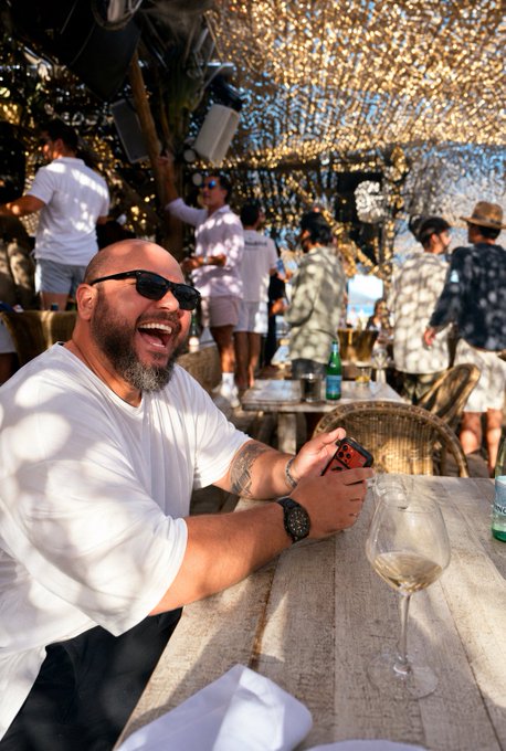 A laughing bearded man wearing sunglasses and a white long-sleeve shirt sits at an outdoor table holding a phone, with a glass of white wine and a lively crowd visible under a tree in a sunlit beachside setting.