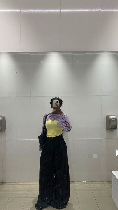 A mirror selfie of a person wearing a purple mesh long-sleeve top over a yellow bandeau, paired with sparkly black wide-leg pants, standing in a modern white-tiled restroom.