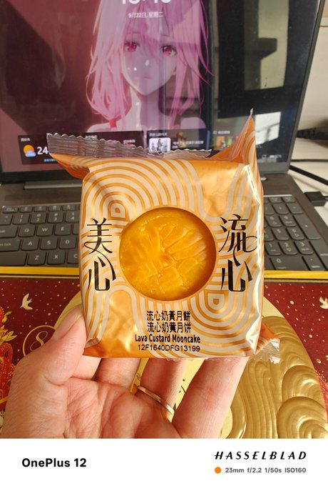 A photo taken on a OnePlus 12 showing a hand holding a 'Lava Custard Mooncake' (流心奶黄月饼) package in front of a laptop displaying an anime character with pink hair.