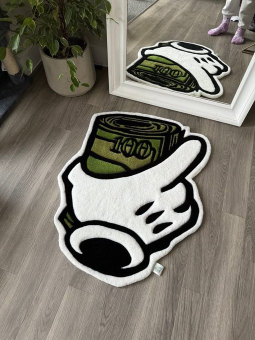 A photograph showing a white rug on a wooden floor, designed to look like a mug of coffee or a skull with a roll of green money (a $100 bill) coming out of it, with a mirror reflecting the design and a person's feet in pink socks visible.