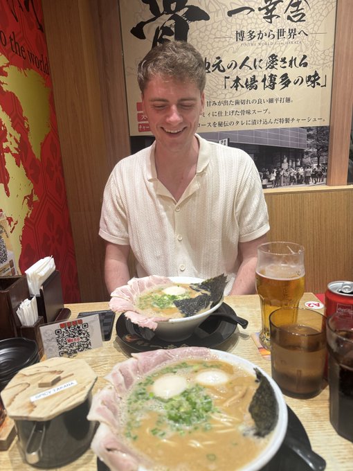 A young man smiling in a Japanese ramen restaurant (一蘭/Ichiran) sitting in front of two bowls of ramen with chashu pork and eggs, with beer and soda glasses beside the meal.