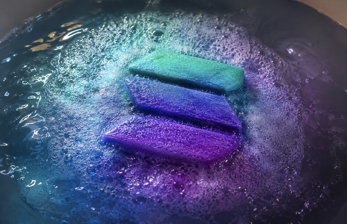 Three bath fizzers in green, blue, and purple dissolve in a dark bath, creating a swirling galaxy of vibrant bubbles and color that visually represents the speculative surge of Solana to $1003. The image's bold, otherworldly color palette evokes the explosive excitement and cosmic optimism surrounding the price prediction.