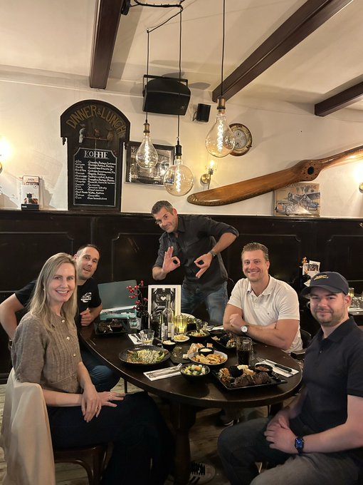 Five people smiling and posing around a table full of food and drinks in a pub setting, with one man standing and making hand gestures; signs on the wall read "DINNER & LUNCH" and "KOHLE".