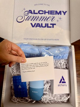 The image shows a person holding up a box containing various items such as a hair dryer, a card, and some other objects. The box has white and blue colors, with the word "Summer" written on it. The person's hand is visible in the foreground of the image, while the box is placed against a wooden background.