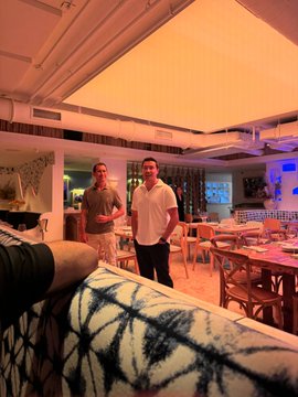 Two men stand in a warmly lit restaurant or lounge area with wooden tables and chairs.