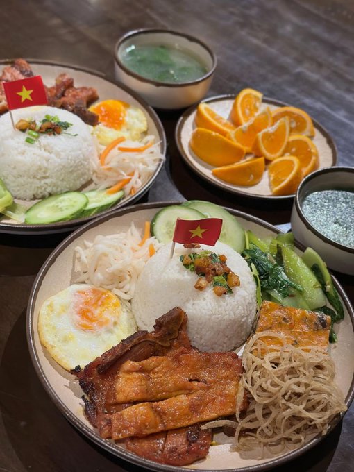 Two plates of Vietnamese broken rice (Cơm tấm) served with grilled pork, fried egg, pickled vegetables, and cucumber, accompanied by a bowl of orange slices and soup, decorated with small Vietnamese flag toothpicks.