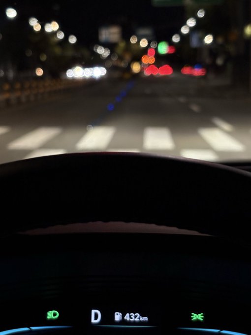 A nighttime view from the driver's seat shows a blurred city street ahead with traffic lights and a crosswalk, while the dashboard displays "D 432km" indicating the drive home. The image pairs with the caption's weary humor about waiting for the crypto bull market to finally arrive—much like waiting out traffic on the evening commute.