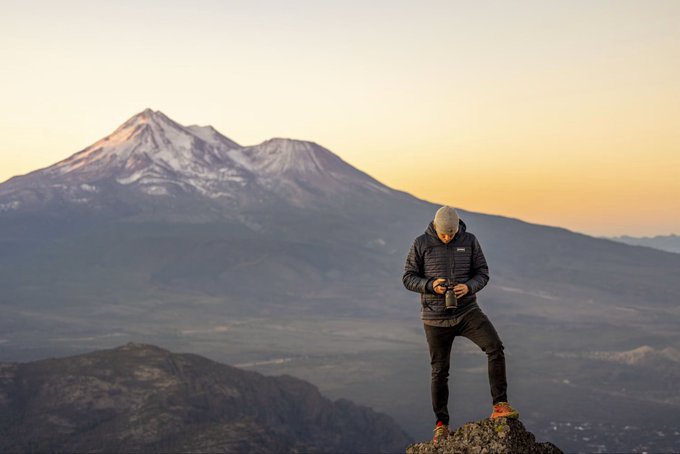 A hiker in a black jacket and beanie stands on a rocky outcrop, pouring from a thermos with a snow-capped mountain (likely Mount Shasta) glowing in the soft golden sunrise behind him. Paired with the caption, the image humorously depicts a peaceful escape to the mountains as an ideal, scenic setting for checking trading charts over the weekend.