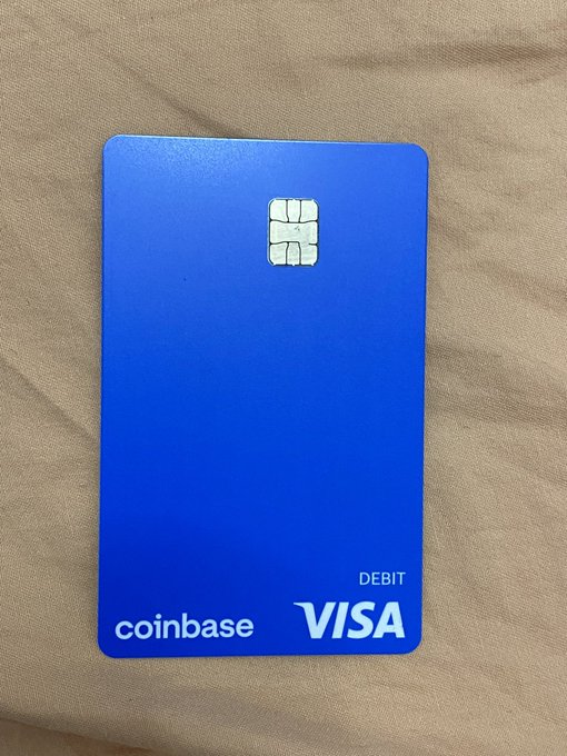 A blue Coinbase Visa debit card is displayed on a beige fabric background, featuring an EMV chip, the "coinbase" logo, "VISA" branding, and "DEBIT" text. The card visually reinforces the post's claim about Coinbase's legitimacy as a major, publicly-backed financial platform by showcasing its branded payment product.
