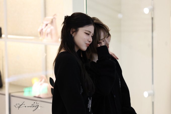 Two young women in black outfits embrace closely in a softly lit room with shelved handbags in the background, with a 'kmj' watermark visible.