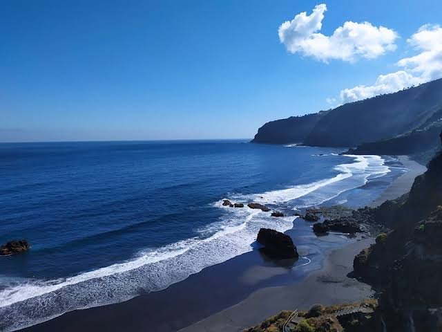 A scenic coastal view showing a beach with waves crashing against the shore, dark cliffs on the right, and a clear blue sky with scattered clouds.