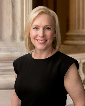 The image shows Senator Kirsten Gillibrand, a blonde woman wearing a black dress, posing for an official portrait against a marble columned background. This relates to the post as she is one of the seven Democratic senators who jointly released a statement reaffirming their commitment to passing the CLARITY Act following a recent setback.