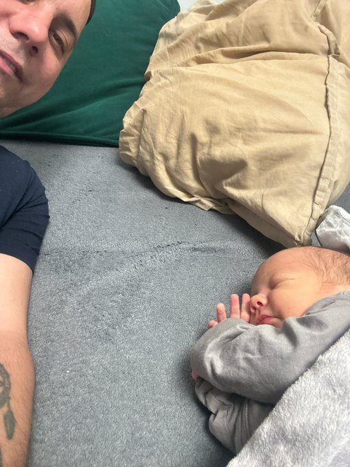 An intimate overhead selfie of a man lying next to a sleeping newborn baby wrapped in grey clothing on a bed with a green pillow.