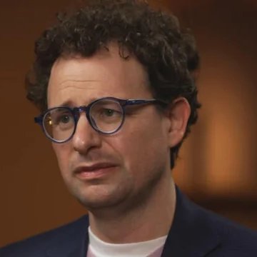 A close-up portrait of a man with curly brown hair wearing blue-rimmed glasses and a dark blazer over a white shirt.