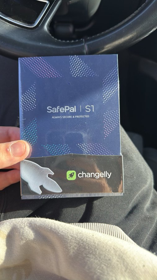 A hand holds a SafePal S1 hardware wallet box, with the Changelly logo visible on the side, while sitting inside a vehicle.