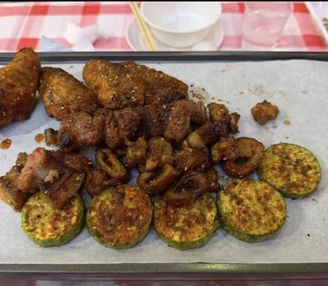 A tray of grilled food including seasoned meat skewers, dark glazed pieces, and sliced zucchini served on parchment paper over a red-checkered tablecloth.