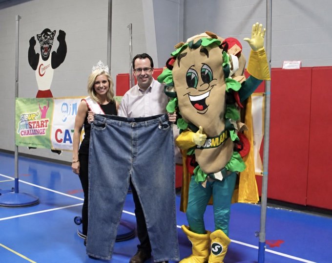 The image features two people, one man and one woman, standing next to a large sandwich mascot costume that appears to be made of lettuce and tomato. The man is holding up a pair of jeans, while the woman stands beside him. They are both smiling for the camera. In the background, there is a banner with the words "smart start challenge" written on it.