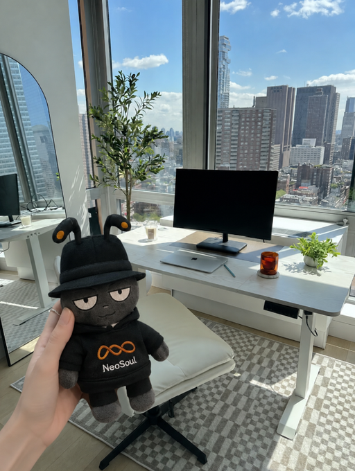 A hand holding a black NeoSoul plush toy wearing a hat with antennae, positioned in front of a desk setup with a monitor and laptop, with a city skyline visible through a large window.