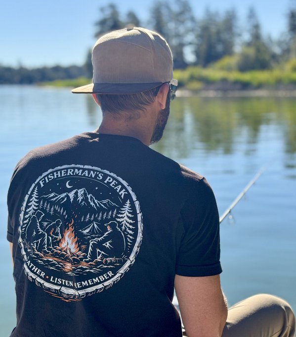 A man wearing a beige cap and a black t-shirt with the text 'FISHERMAN'S PEAK GATHER LISTEN REMEMBER' sitting by a lake, fishing.