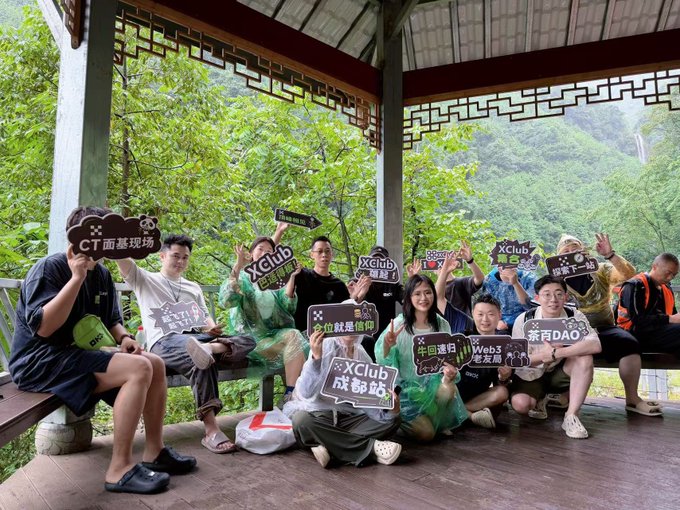 A group of tired but cheerful hikers pose together under a traditional Chinese pavilion, holding up signs reading "XClub 成都站" (XClub Chengdu) and other OKX-branded markers like "CT 面基现场" and "回家DAO." The image captures the exhausted yet fun atmosphere of an OKX XClub community hiking event, with participants flashing peace signs and smiling in their rain ponchos after a rainy mountain trek.