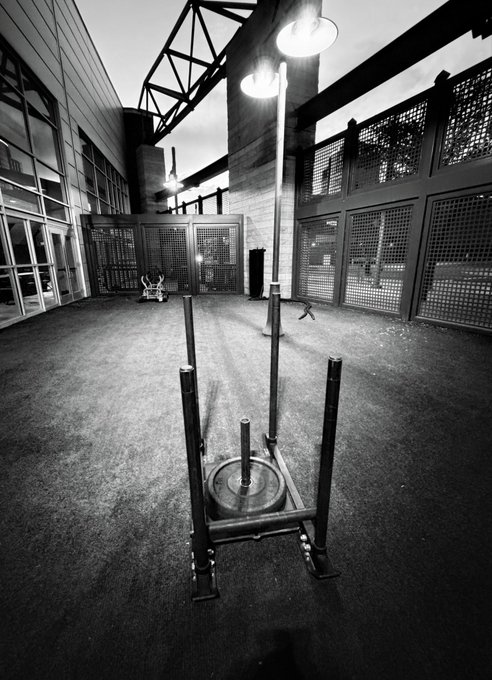 A black-and-white photograph of an empty gym featuring a barbell with weights in the foreground, a tall lamp post in the center, and caged workout areas along the walls.