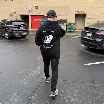 A man wearing a black 'BAPE' hoodie with a panda logo walks through a parking lot past parked cars.