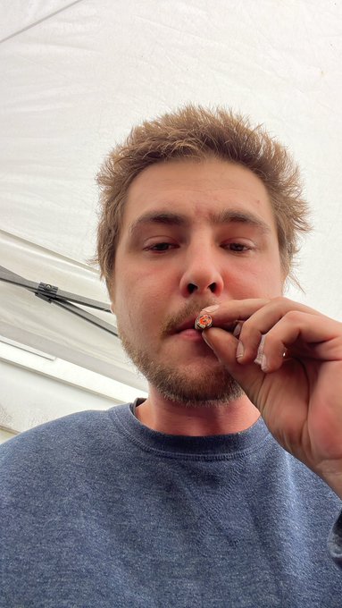 Close-up selfie of a man with tousled brown hair and a light beard, wearing a gray sweater and holding what appears to be a cigarette or small object near his lips.