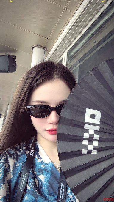 A young woman with dark hair wears black sunglasses and holds a black folding fan with the "DJI" logo, photographed in what appears to be an indoor event space. The "F1 Chance Encounter" caption suggests an impromptu selfie at an F1 racing event.