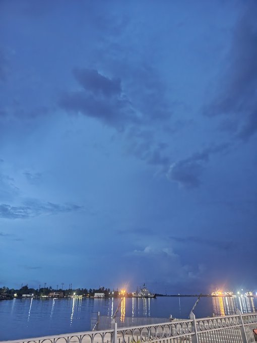 A twilight scene shows a body of water with city lights twinkling in the distance. A white railing is visible in the foreground.