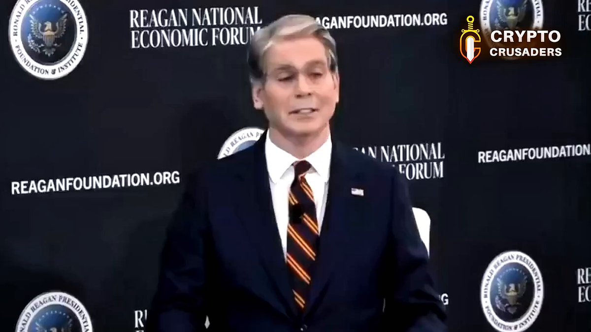 The image features a man dressed in a suit and tie standing against a black background with white text, including logos for the Reagan National Economic Forum and the U.S Treasury Department. The man appears to be speaking or giving a speech at an event, possibly related to cryptocurrency. The caption of the image suggests that there is some sort of discussion about crypto-related matters taking place in front of these organizations.