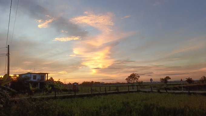 A scenic photograph of a sunset over a grassy field, with a small house and trees silhouetted against the cloudy orange sky.