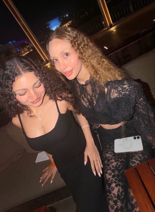 Two young women posing together at night, both wearing black outfits - one in a black tank top and the other in a black lace top and skirt with a white smartphone in her pocket.