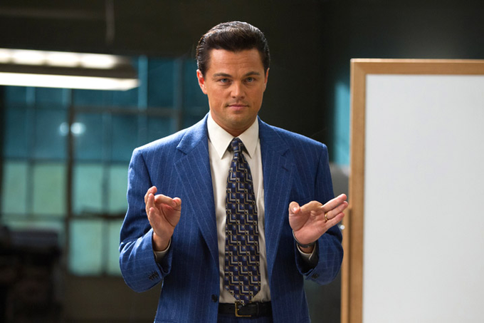 Leonardo DiCaprio as Jordan Belfort in a blue pinstripe suit and patterned tie, gesturing with both hands in what appears to be a scene from The Wolf of Wall Street.