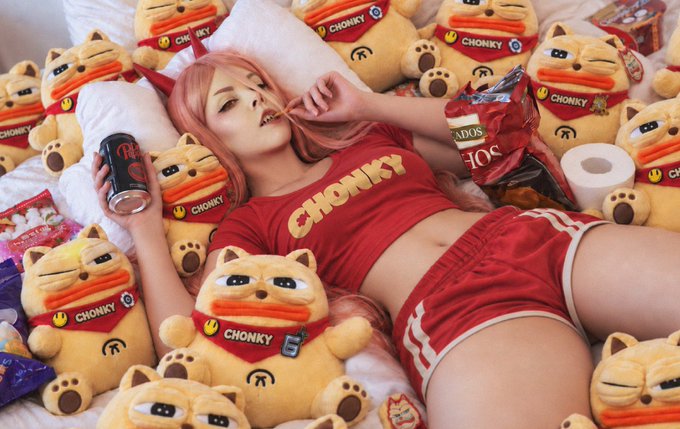A cosplayer with pink hair lies on a bed surrounded by plush toys wearing red 'CHONKY' bandanas, wearing a matching red 'CHONKY' shirt and shorts, and holding a snack and a drink.