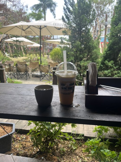 An iced Vietnamese coffee in a plastic cup with a domed lid and straw sits on a dark wooden table alongside a small dark cup and a napkin holder, with text reading "CÀ PHÊ THÁI LAN" visible on the cup, set against a lush garden patio with umbrellas and chairs in the background.

The caption and image appear loosely connected thematically—both suggest something holding up well amid a broader decline—though the outdoor coffee scene doesn't directly illustrate the Bitcoin rally discussion.