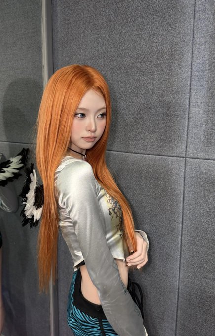 A person with long orange hair posing against a gray tiled wall, wearing a silver/gray satin long-sleeve top and patterned pants.