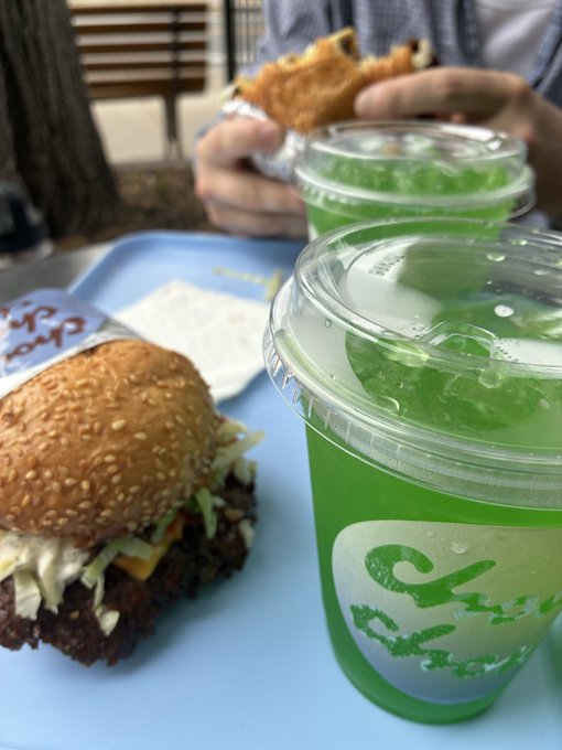 A meal at a casual restaurant featuring a burger with sesame seed bun, green iced drinks in plastic cups with 'Goya' branding, and a person eating in the background.