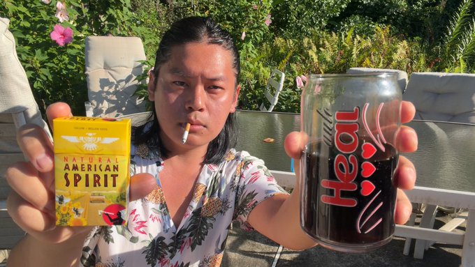 A young man with long dark hair smokes a cigarette while holding up a yellow pack of "Natural American Spirit" cigarettes and a "Heal" branded glass, sitting outdoors in a sunny garden setting that visually reinforces the post's theme of contemplation and smoking.