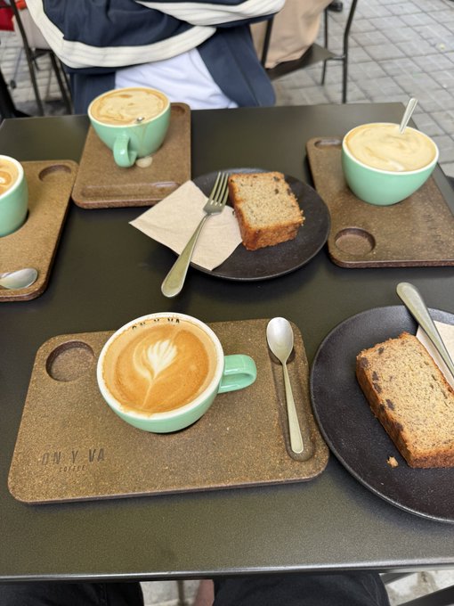 A coffee table with three mint green cups of latte art on cork coasters branded "MY VVA COFFEE," alongside slices of cake on dark plates.