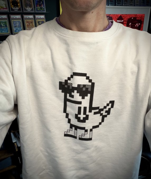 A person wearing a cream-colored sweatshirt featuring a pixel-art graphic of a small character wearing sunglasses.